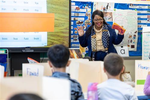 Dr. Natalyn Gibbs standing in front of a classroom, waiving to a group of elementary students.
